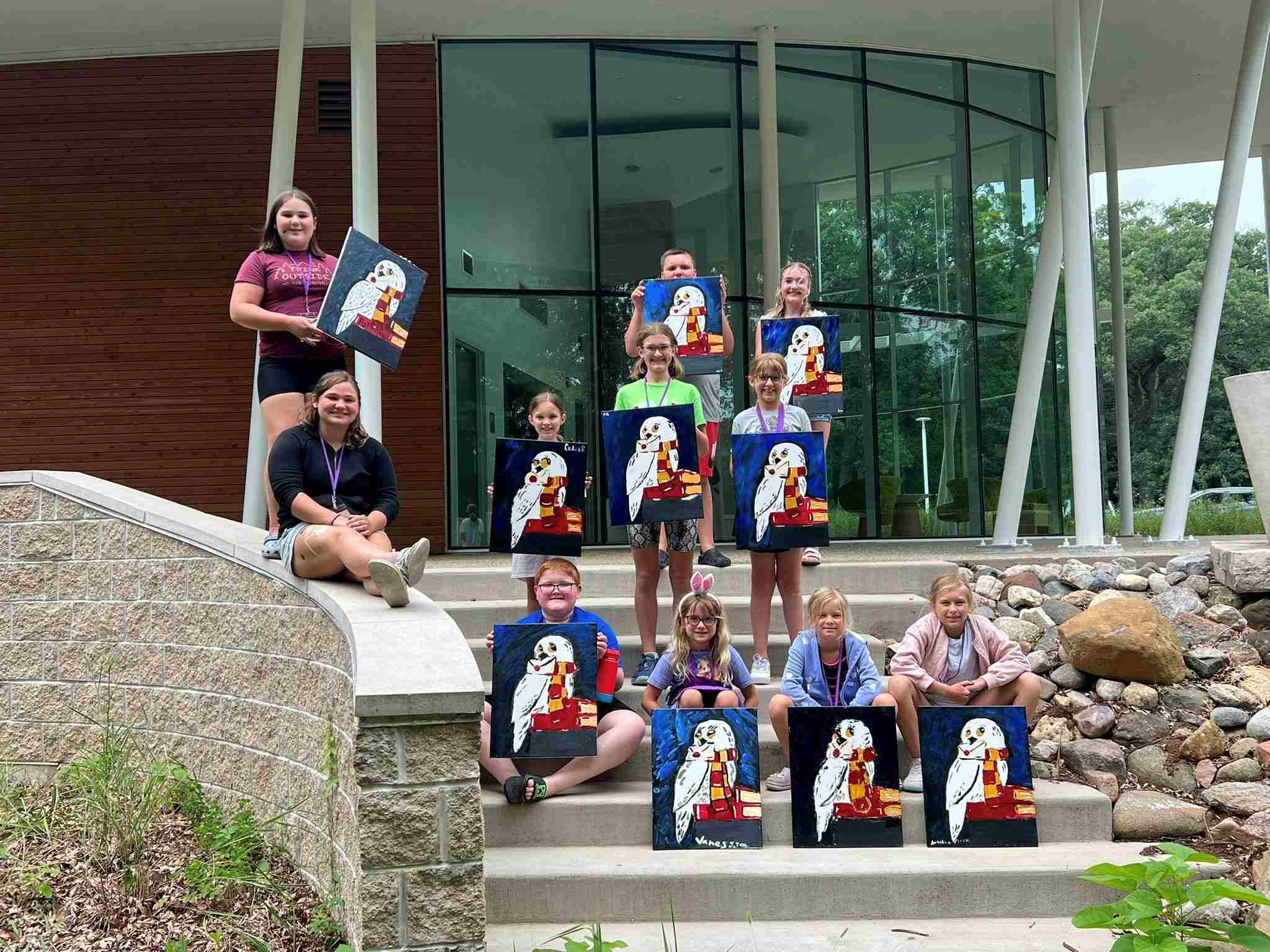 Multiple summer camp students at Nell's Woodland, standing on steps, holding canvas paintings featuring an owl.